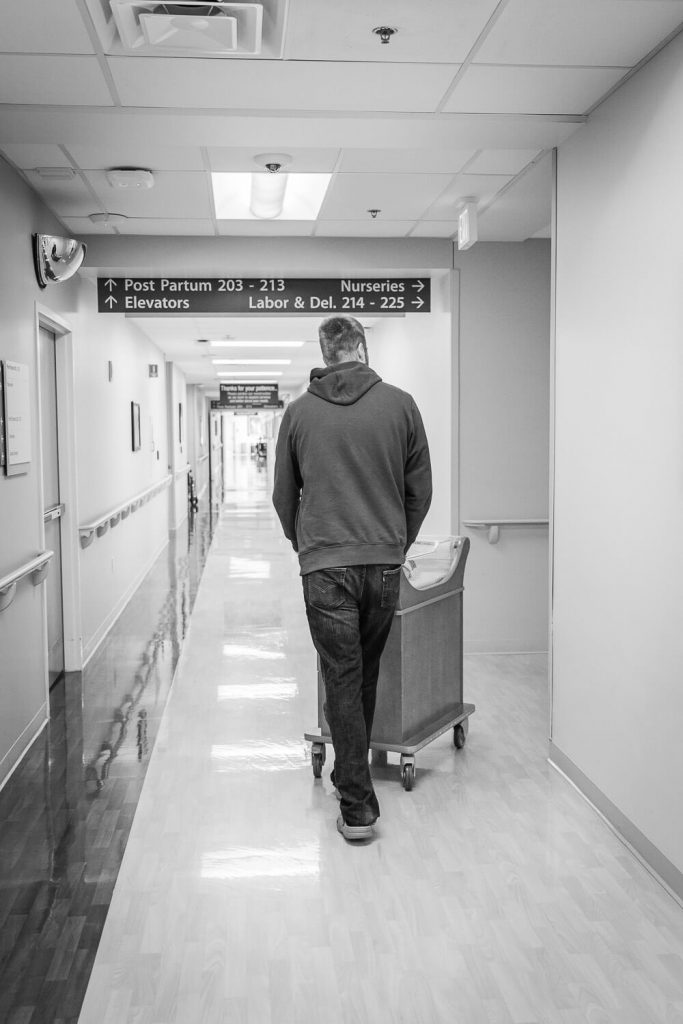 black and white dad pushing bassinet through hospital hallway