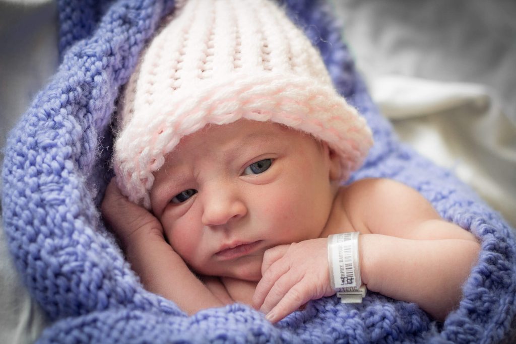 newborn girl wearing white crochet hat wrapped in a purple crochet blanket staring at the camera