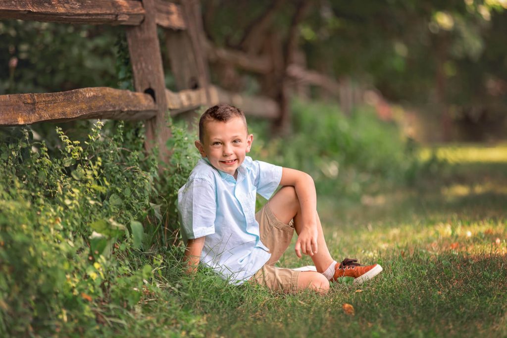 young boy sitting leaned against a wooden fence in a tall grass field