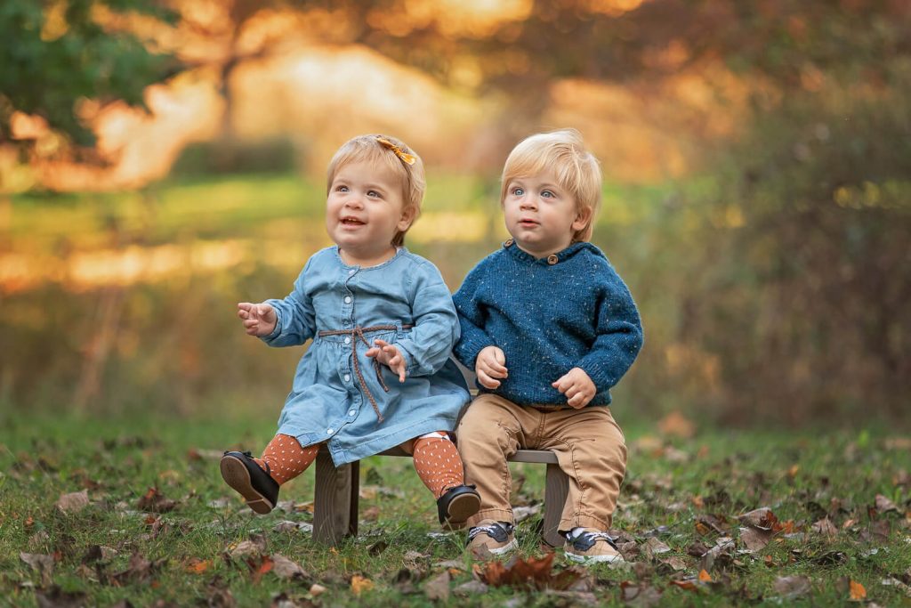 twin 1 year old siblings sitting on a bench under autumn trees