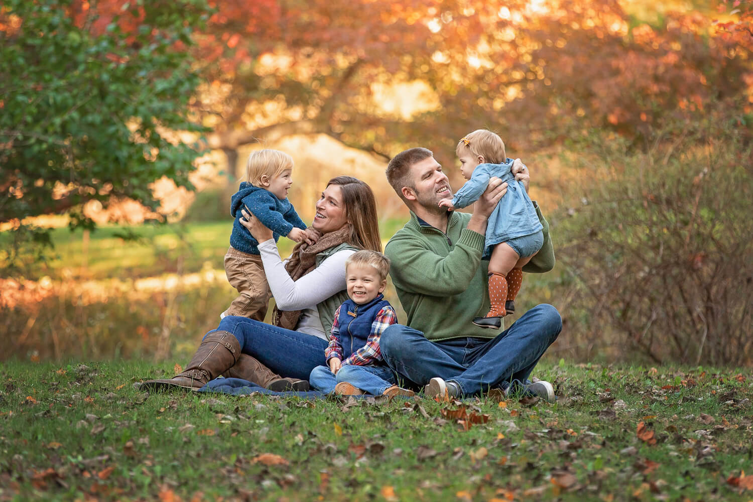 Family of 5 interacting on a blanket on the grass surrounded by bright fall leaves
