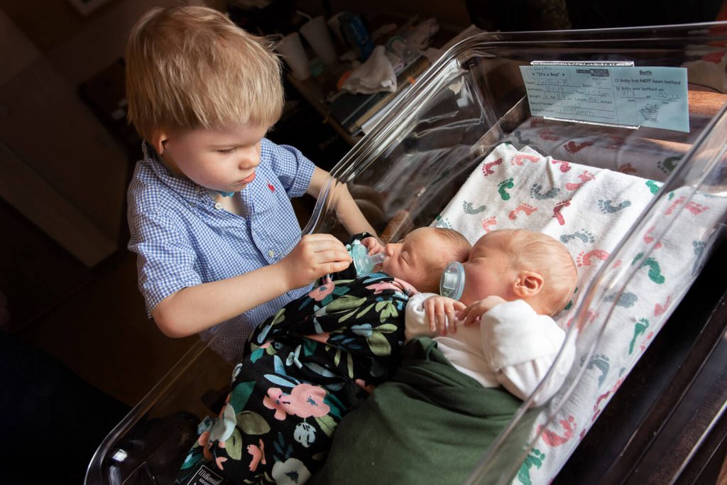 big brother giving a pacifier to one of his newborn twin siblings in a hospital bassinet