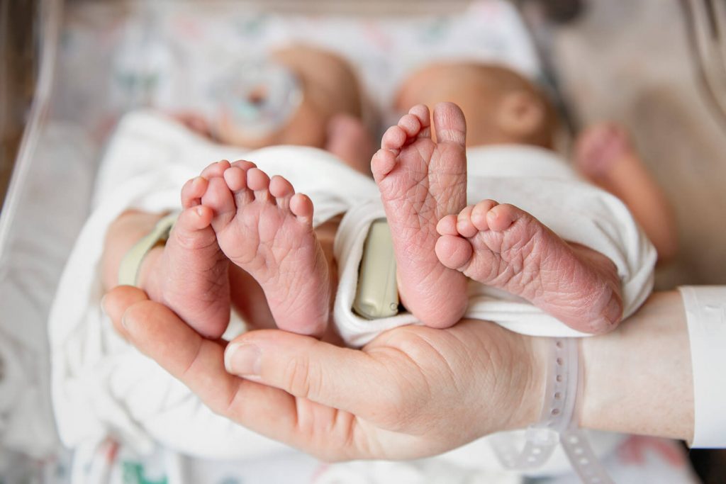 mom holding the feet of twin newborns in the hospital bassinet