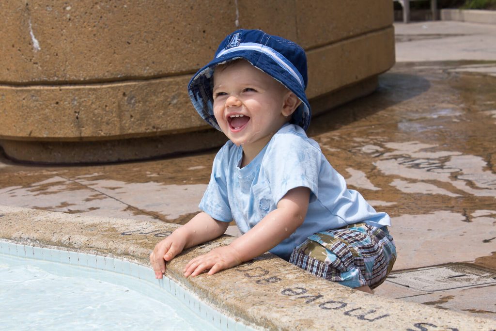 baby boy wearing a blue fishing hat smiling and splashing in water at Meijer Gardens