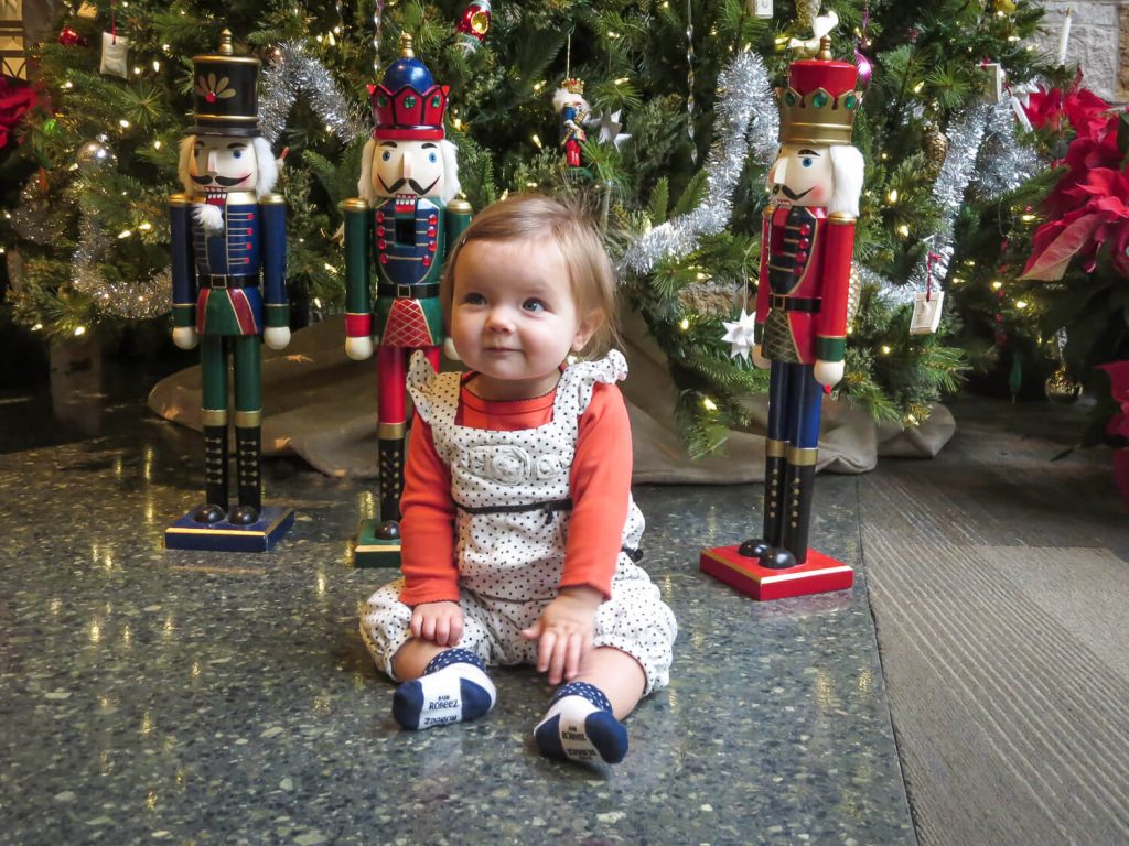 baby girl sitting on the floor in front of nutcrackers and Christmas trees at Meijer Gardens