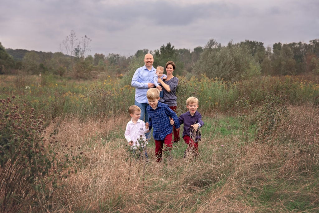 Mom, Dad and baby boy standing in the background while 3 young brothers hold hands and race towards the camera through a tall grass field at Roselle Park