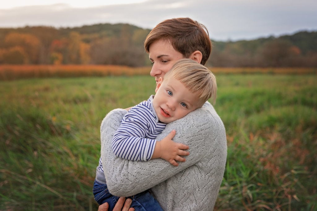 Baby boy leaning his head on mom's shoulder in a tall grass field at sunset in Roselle Park