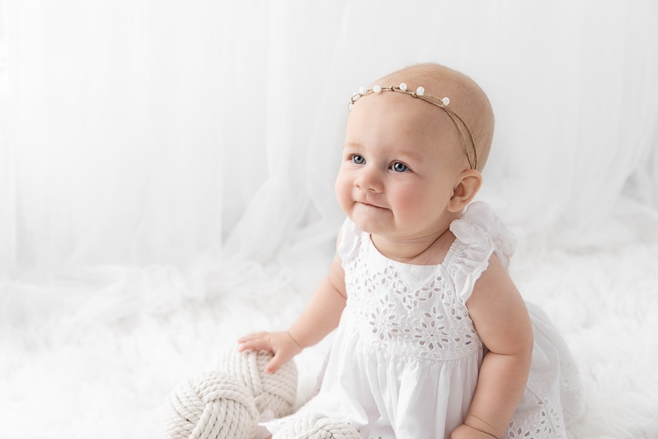 Backlit 6 month baby girl wearing a white dress sitting on a white fur in front of sheer white curtains