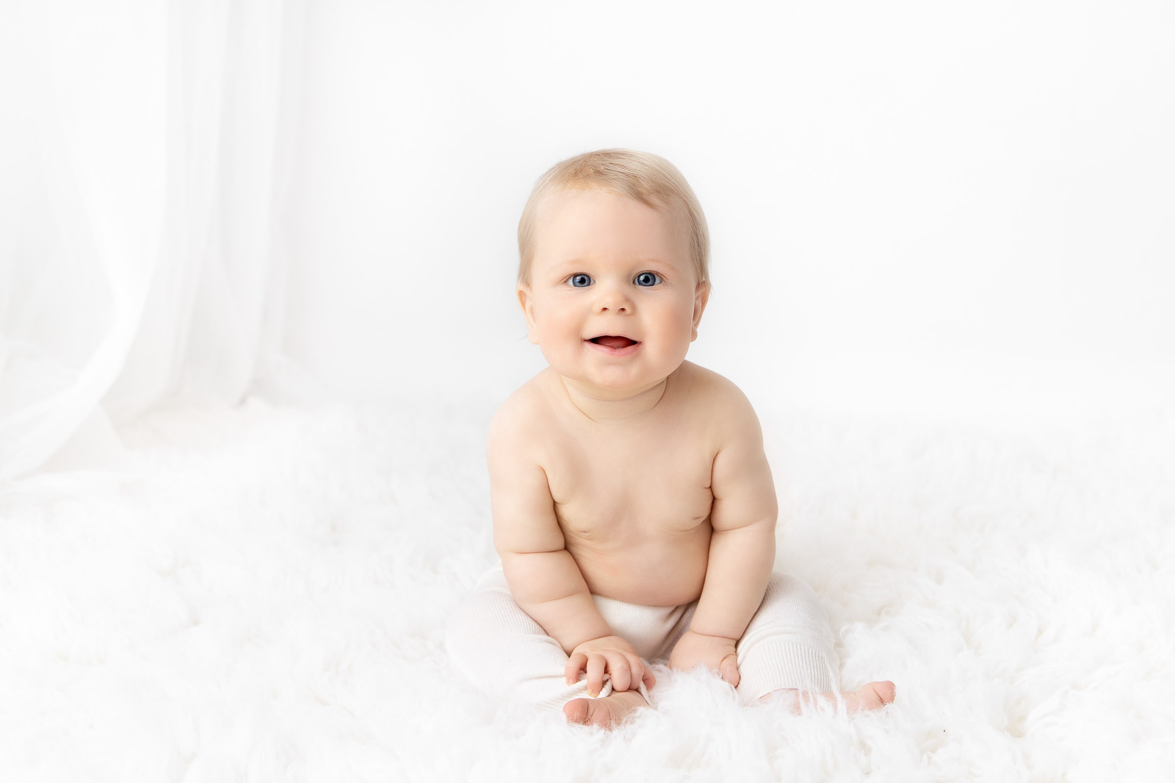 6 month old baby boy wearing white pants sitting on a white fur rug in a white studio smiling at camera