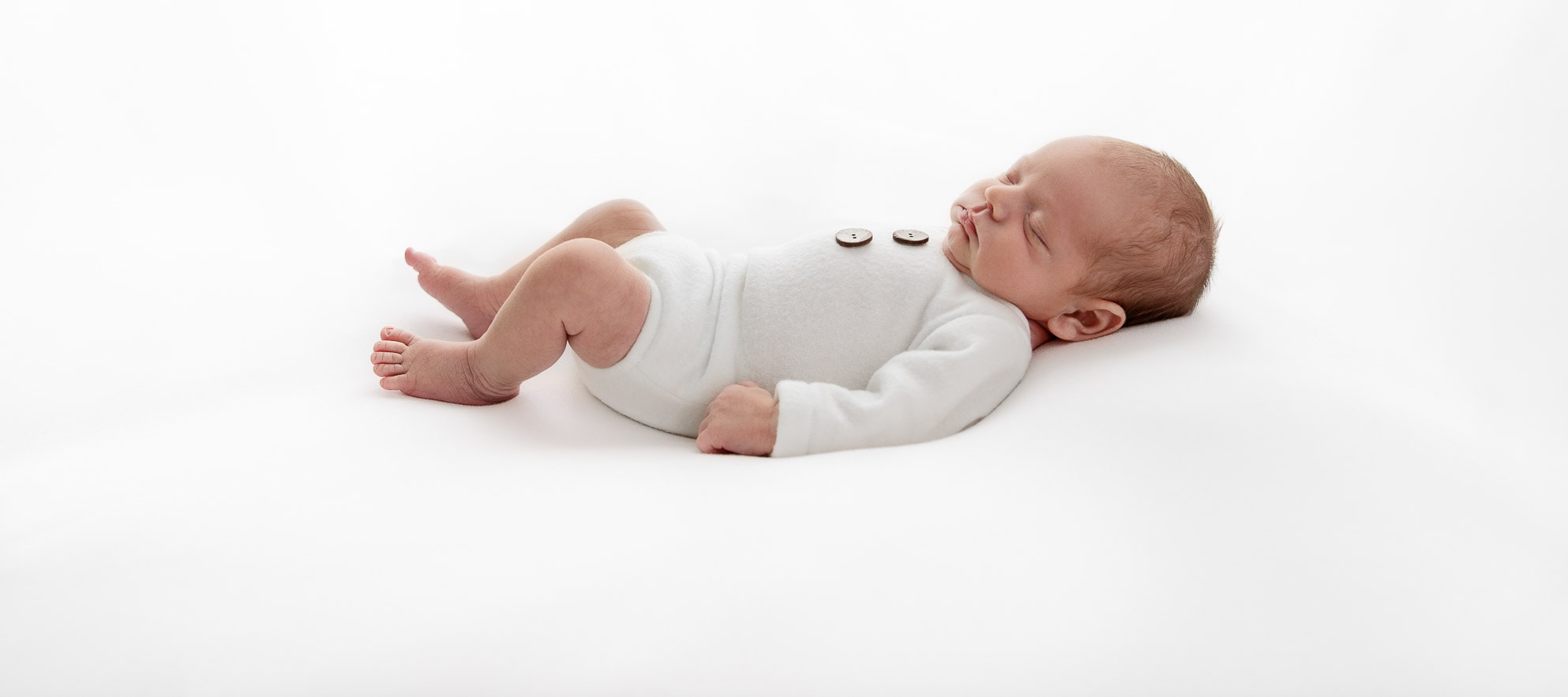 Backlit newborn baby boy sleeping on his back wearing a white romper on a white background