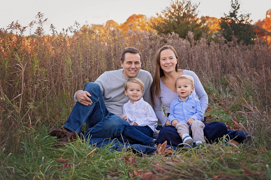 Family of 4 sitting together on a blanket in the middle of a tall grass field at sunset