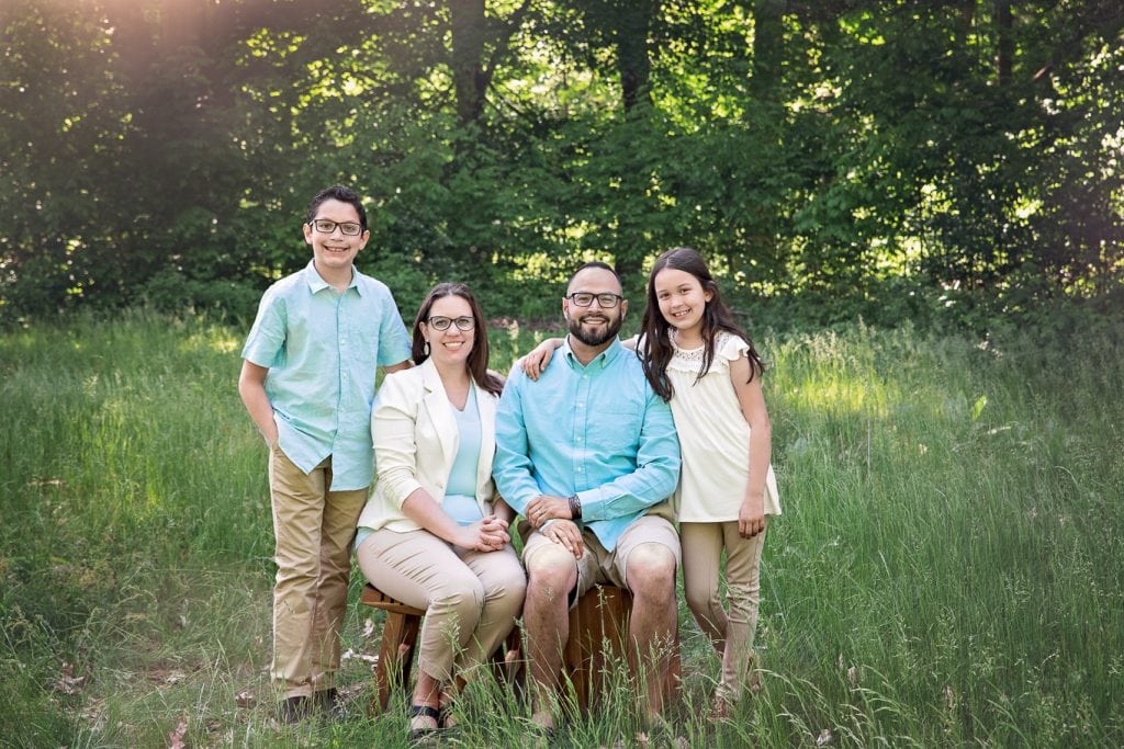 Family of 4 posed together in a green grass field with sunflare shining through green trees in the background