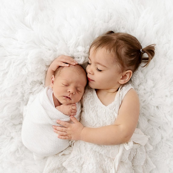 toddler sister with eyes closed holding sleeping baby brother on white fur