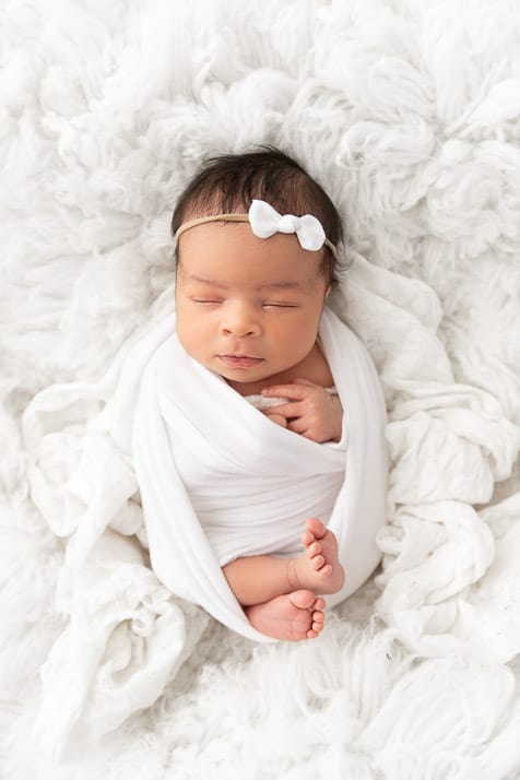 newborn baby girl sleeping on her back snuggled in a white fur wearing a white wrap and white bow