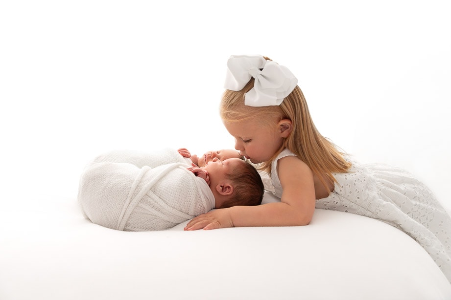 backlit white image of toddler girl kissing her twin siblings on the head