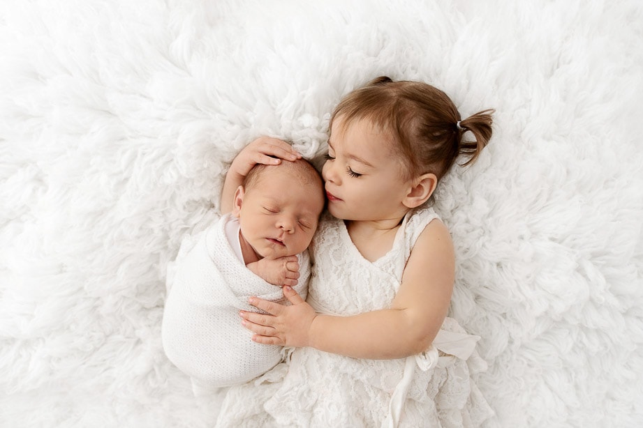 toddler sister with eyes closed holding sleeping baby brother on white fur