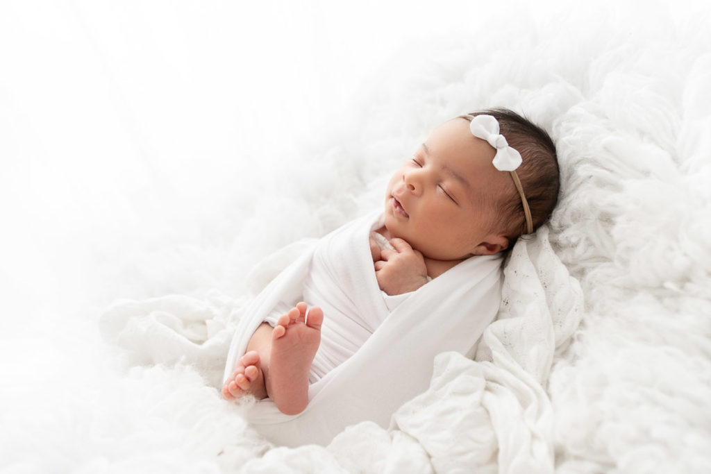Newborn baby girl snuggled in fluffy white fur while wrapped in a white fabric