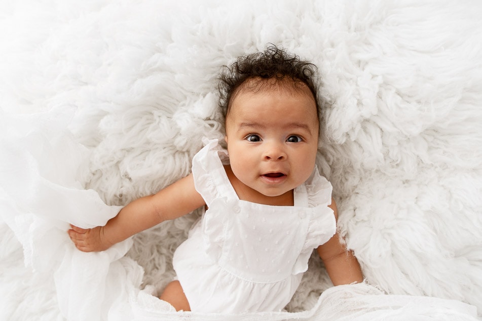 3 month baby girl wearing a white romper laying on a white fur looking up at camera
