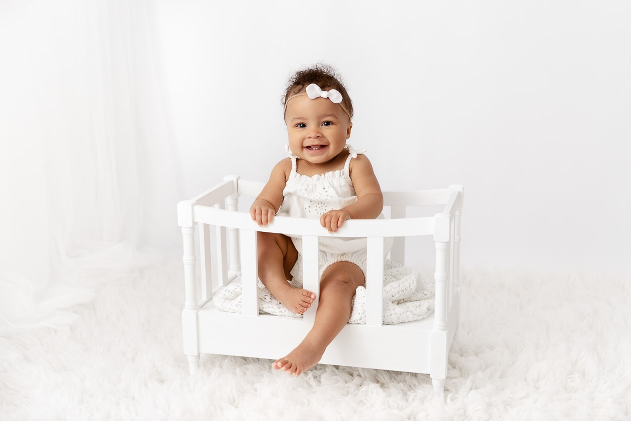 9 month old baby girl wearing a white romper sitting in a small white crib prop on a white flokati fur