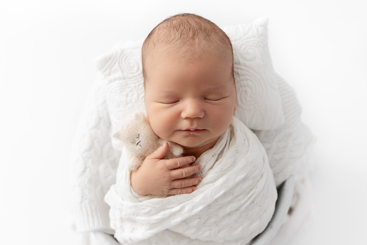 Newborn baby boy wrapped in a white swaddle sitting in a white bucket holding a knit bear prop