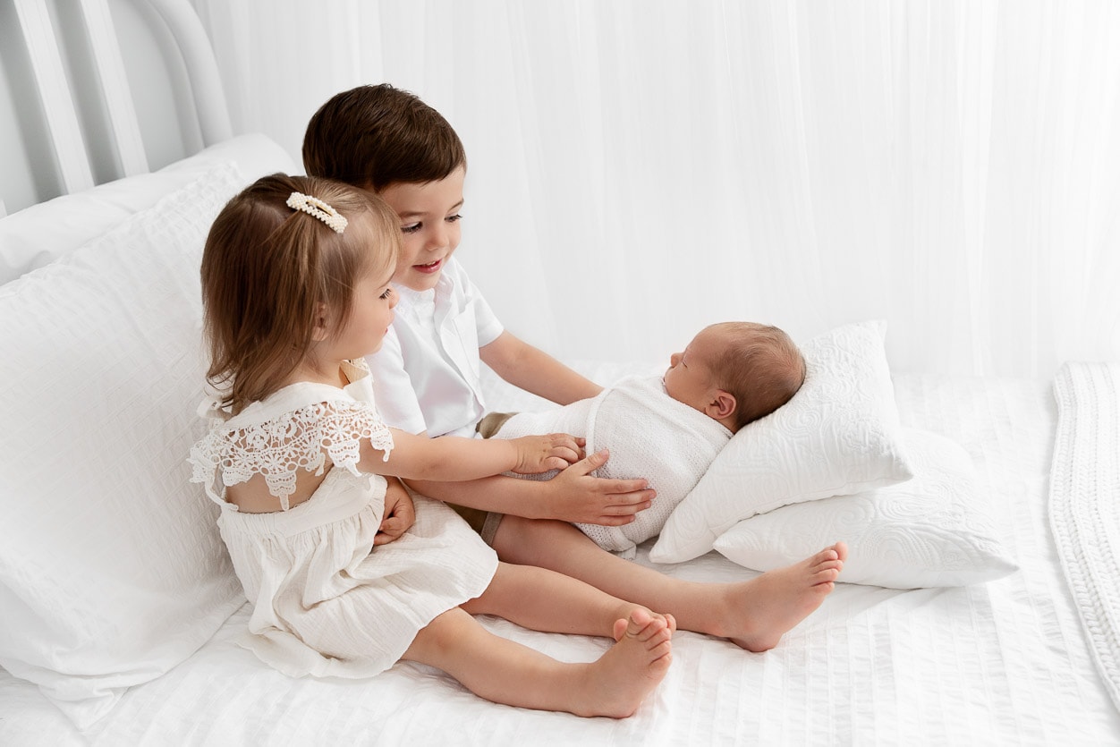 Siblings sitting on a white bed looking down at their newborn baby brother
