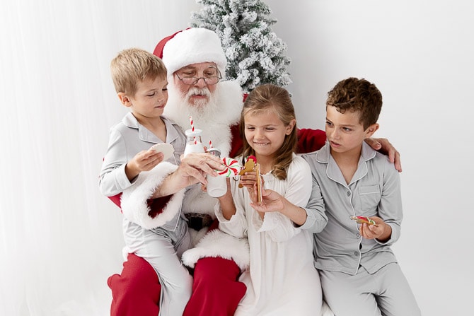 Three young kids sitting with Santa on a bench sharing milk and cookies