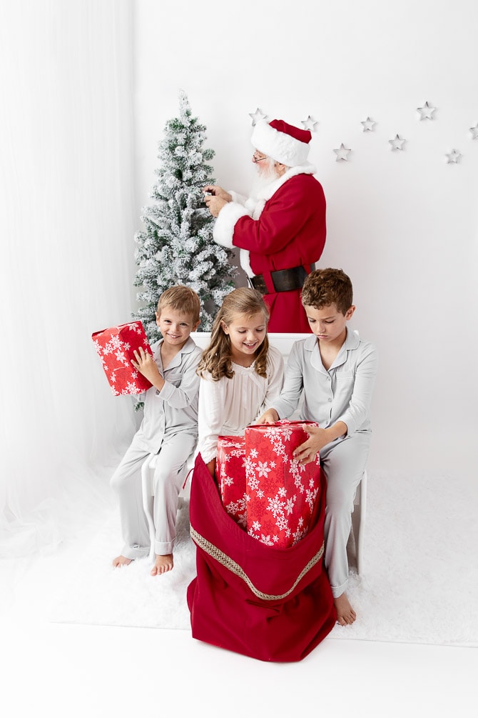 Three young kids sit on a bench looking through a red sack of presents while Santa decorates a tree in the background
