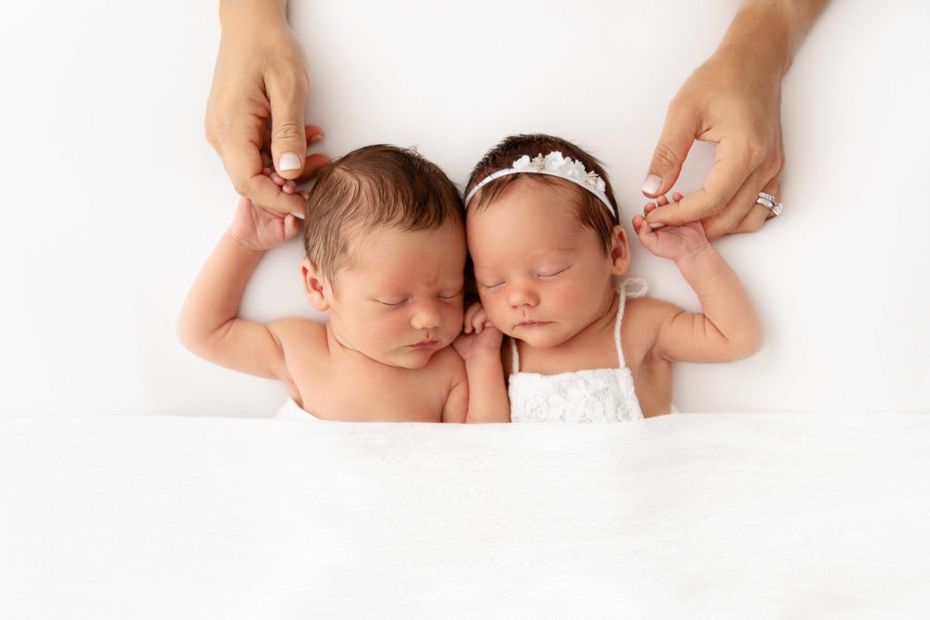 Newborn boy girl twins sleeping on a white bean bag holding mom's hands