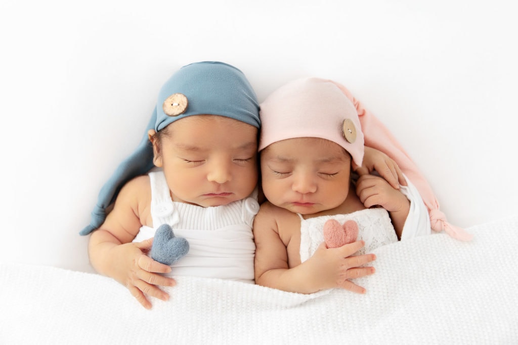 Newborn boy girl twins sleeping with arms around each other wearing sleepy caps and holding felted hearts