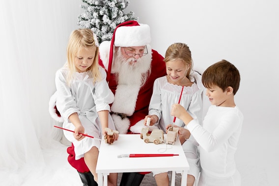 Three kids wearing white pajamas sitting with Santa painting wooden toys
