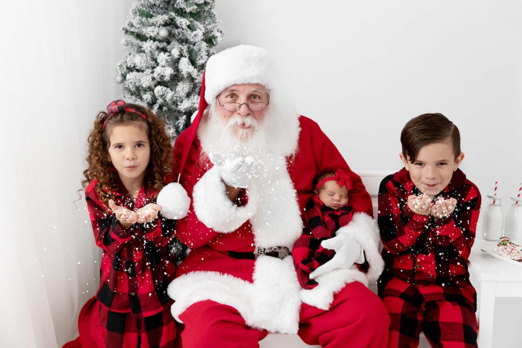 Three children wearing red plaid pajamas blowing snow with Santa during their holiday photo session