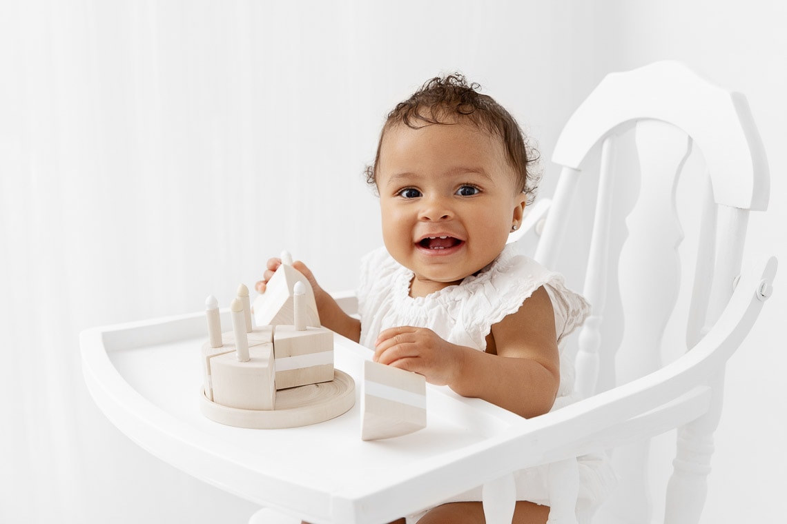 One year old baby girl sitting in a white wood high chair playing with a natural wood cake prop