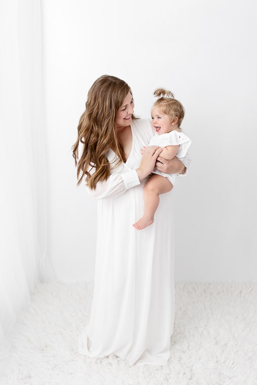 mom holding and playing with one year old baby girl in front of a white sheer curtain
