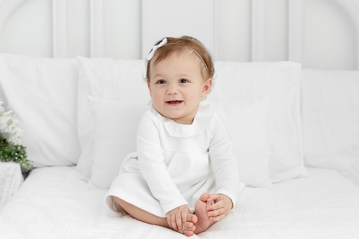 One year old girl sitting and smiling in the middle of a white lifestyle bed