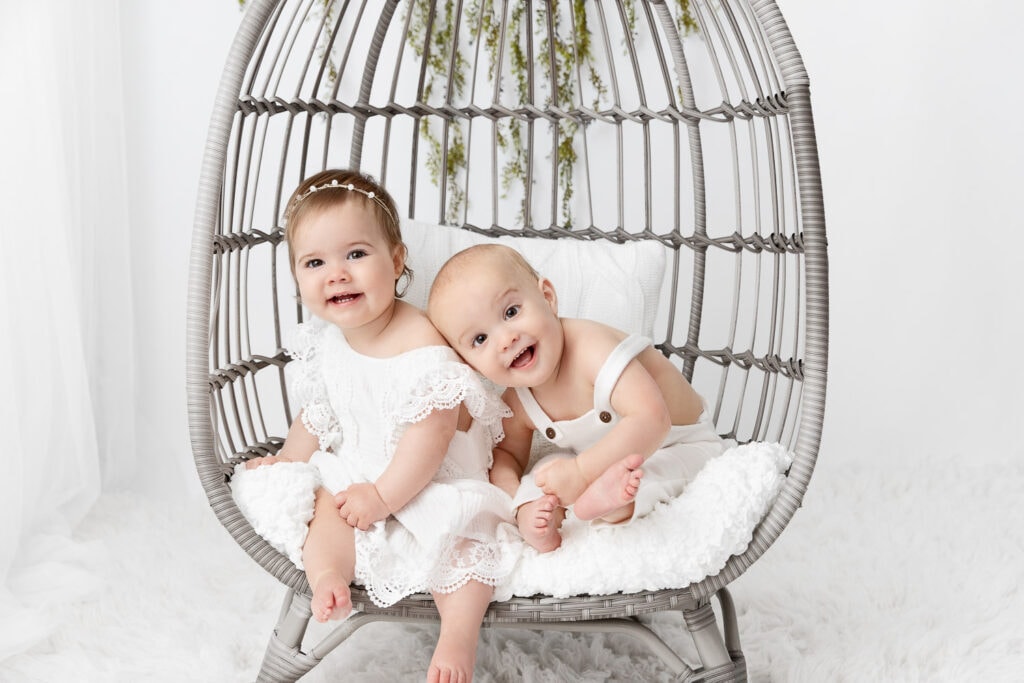 Boy girl one year old twins leaning into each other while sitting in a basket chair in a white studio 