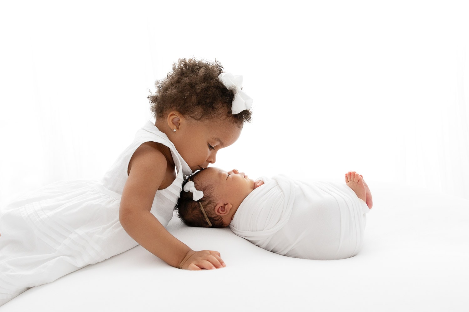 backlit image of toddler girl kissing the top of the head of newborn baby sister