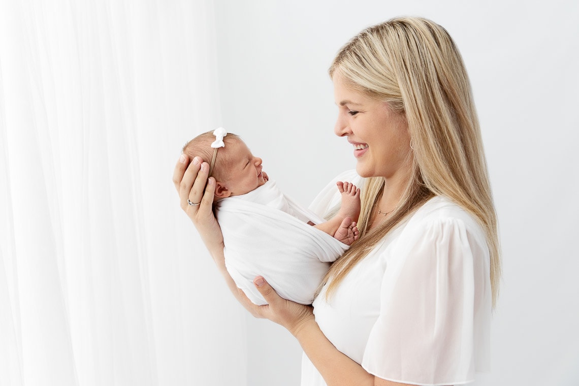 mom holding smiling newborn daughter face to face in front of white curtain