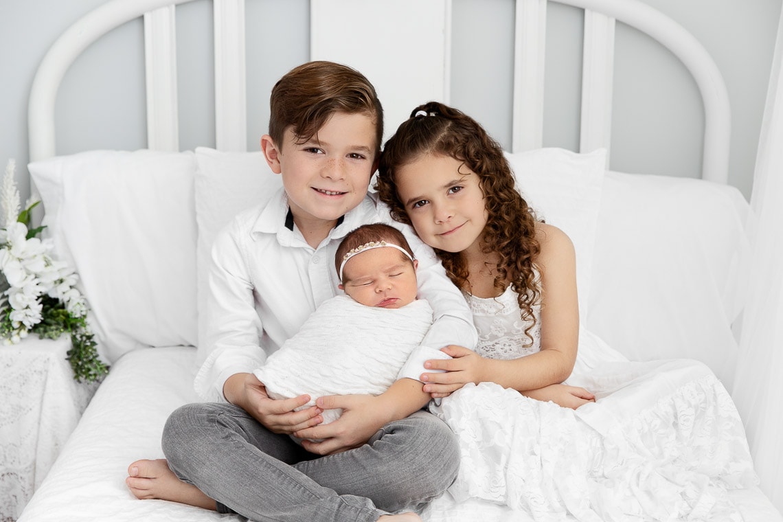 siblings holding newborn baby sister sitting on a bed in a white studio