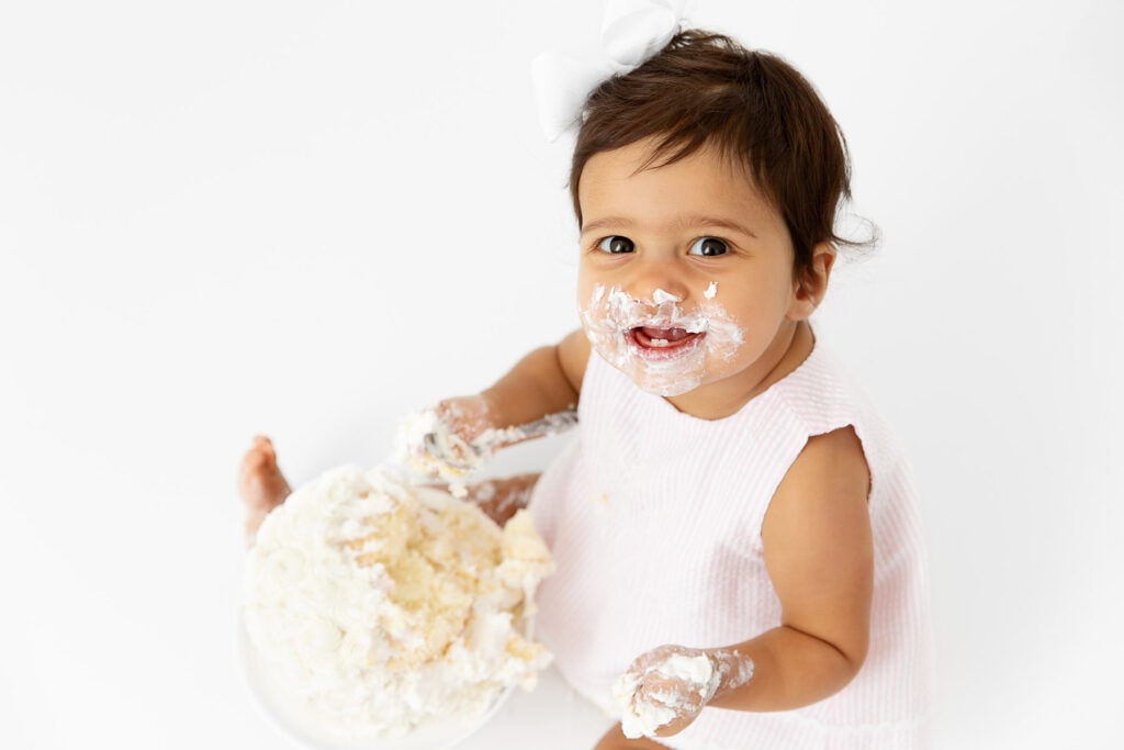 1 year old baby girl smiling up at camera with frosting all over her face with a smash cake