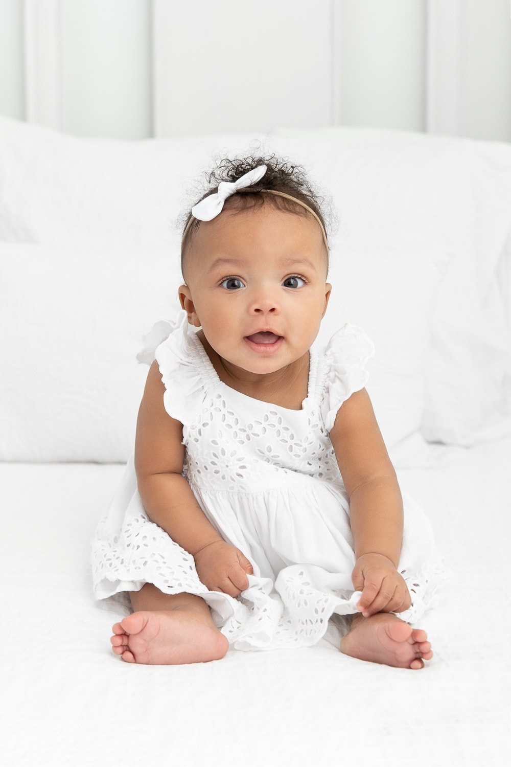 6 month old baby girl siting on lifestyle bed wearing a white eyelet dress with simple white bow