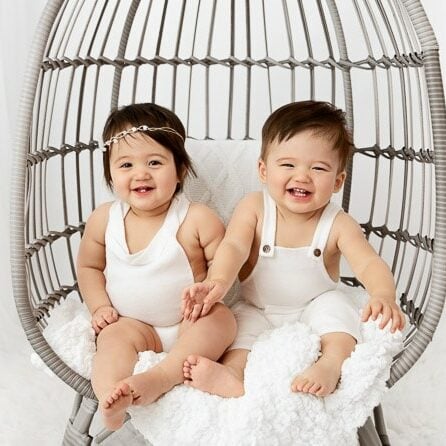 one year old boy girl twins sitting in a gray egg chair together in a white studio