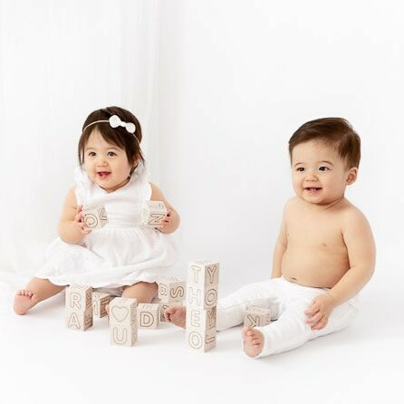 1 year old boy girl twins sitting in front of a white sheer curtain playing with wooden letter blocks
