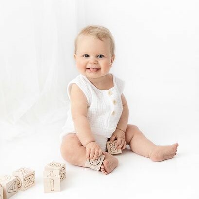6 month baby boy sitting in a white studio in front of white sheer curtains playing with classic wooden blocks