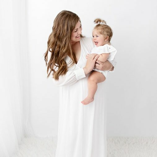 mom holding and playing with one year old baby girl in front of a white sheer curtain