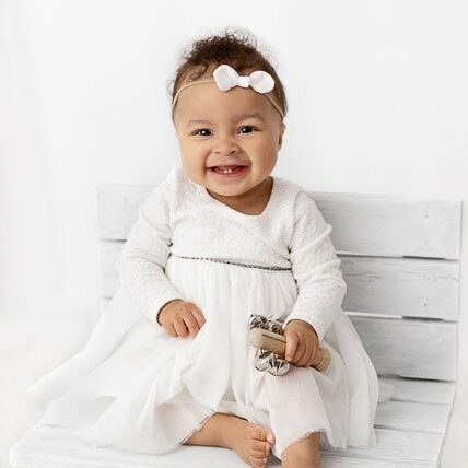 9 month baby girl wearing a white dress sitting on a white bench in front of a white sheer curtain