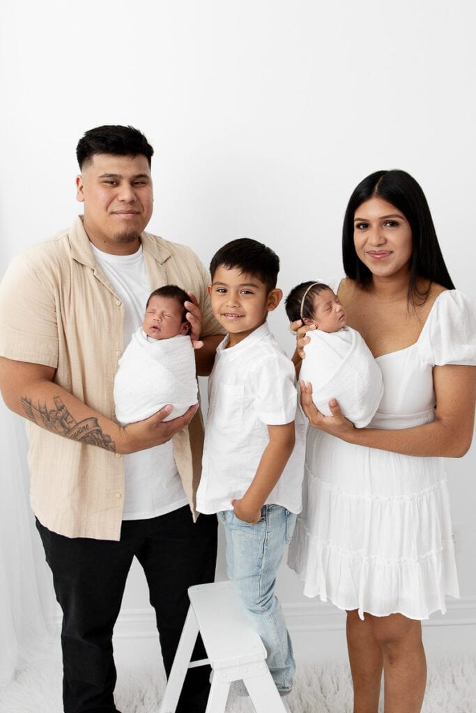 Family of 5 with newborn baby twins and big brother standing together in front of a white wall