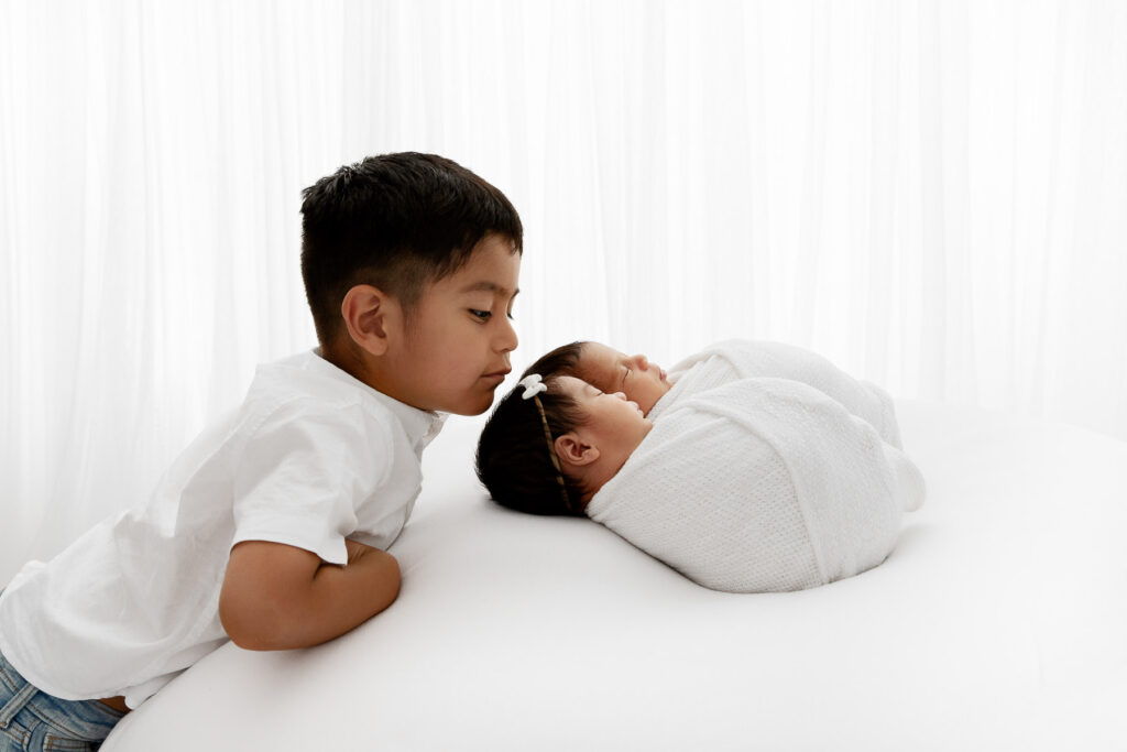 backlit image of big brother gazing at newborn twin siblings who are swaddled on a white beanbag