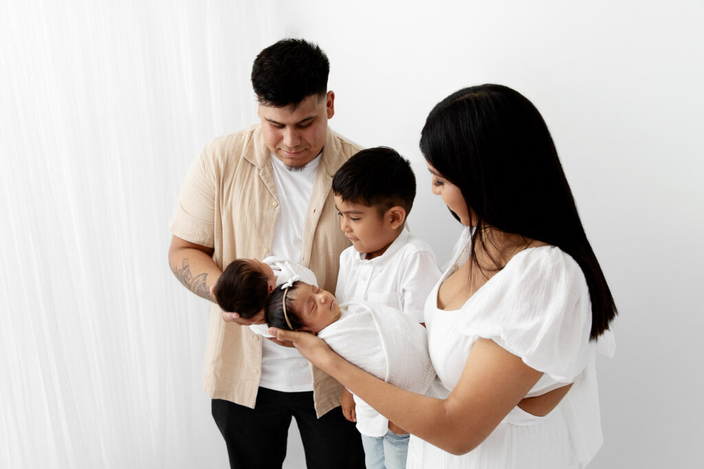 Family of 5 standing in front of white curtain staring down at swaddled newborn baby twins