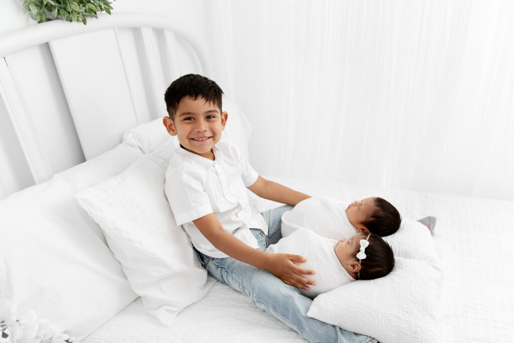 backlit image of big brother holding swaddled newborn twin siblings on white lifestyle bed
