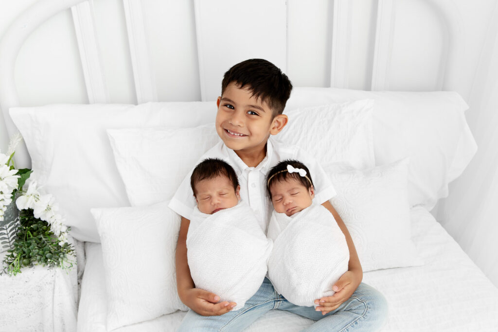 big brother smiling up at camera while holding newborn twin siblings on white lifestyle bed
